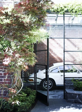 Outdoor view of a white Porsche through a black-framed glass door, with greenery and a brick wall adding to the aesthetic.