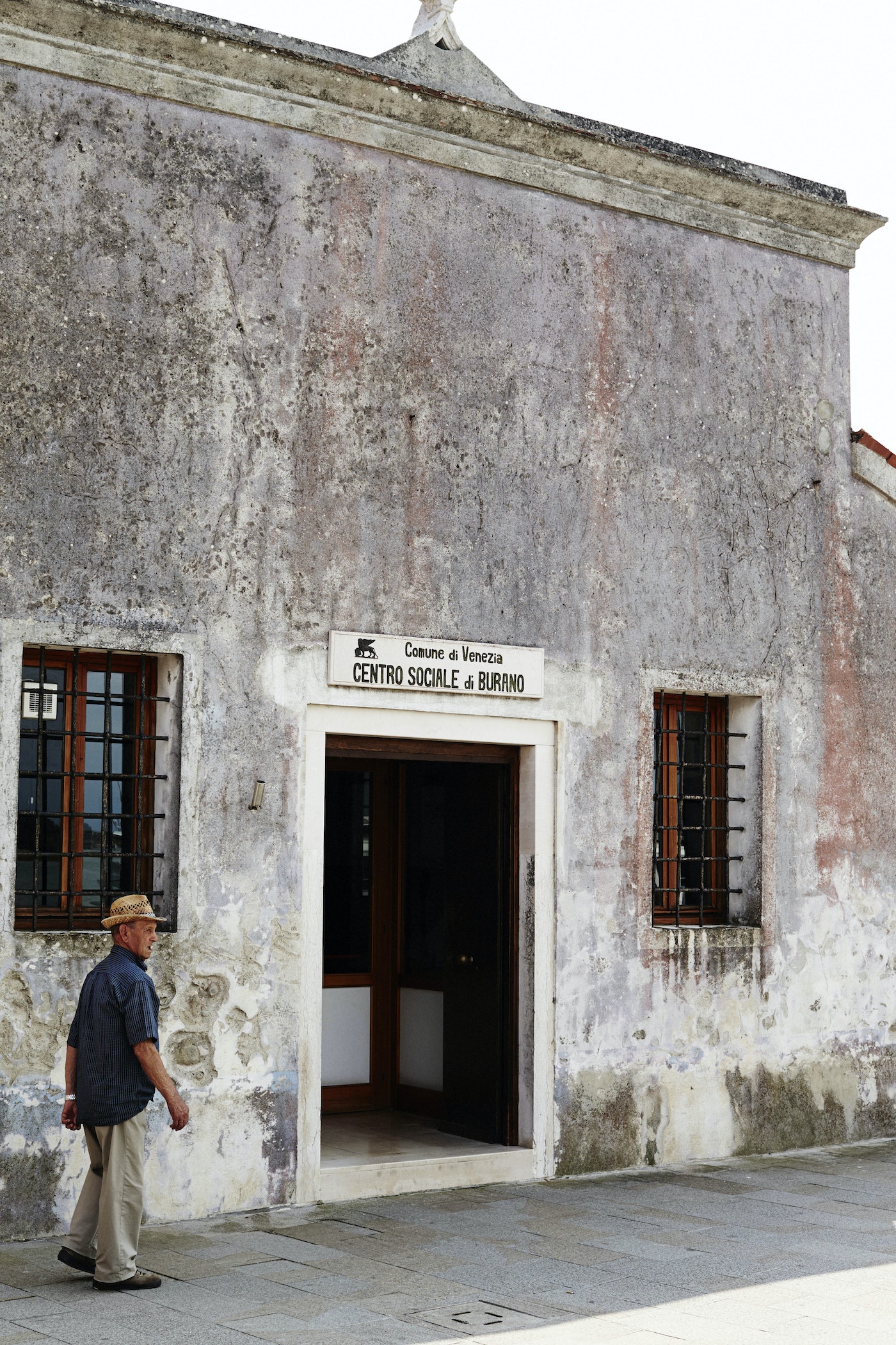 An adult male wearing a hat and casual attire walks past a weathered building with windows and a doorway on a sunny day.