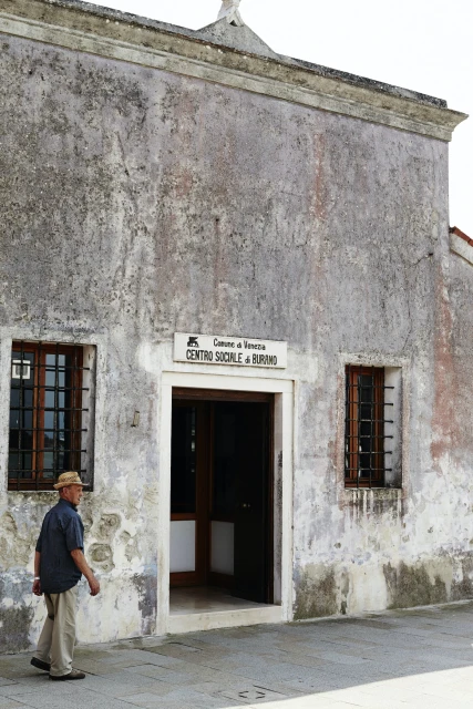 An adult male wearing a hat and casual attire walks past a weathered building with windows and a doorway on a sunny day.