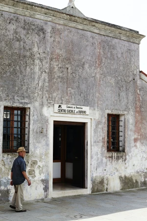 An adult male wearing a hat and casual attire walks past a weathered building with windows and a doorway on a sunny day.