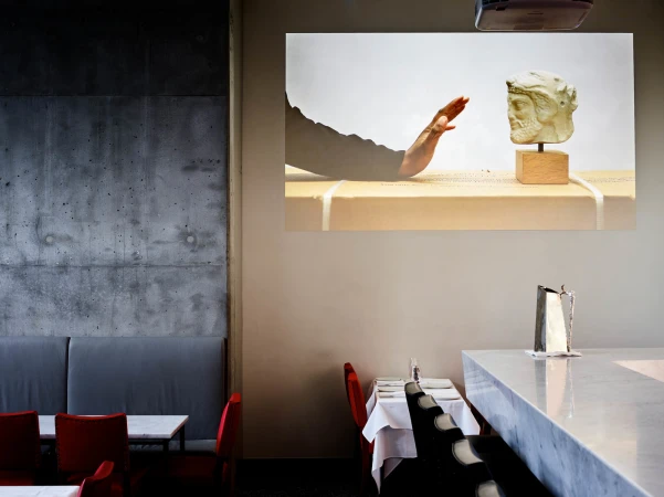 Interior restaurant scene featuring a projected image of a hand reaching toward a stone bust sculpture on a counter.