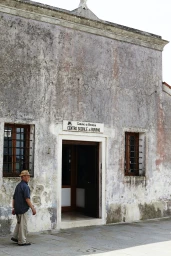 Elderly man wearing a straw hat and striped shirt walking past a weathered stucco building entrance by Mark Roper.