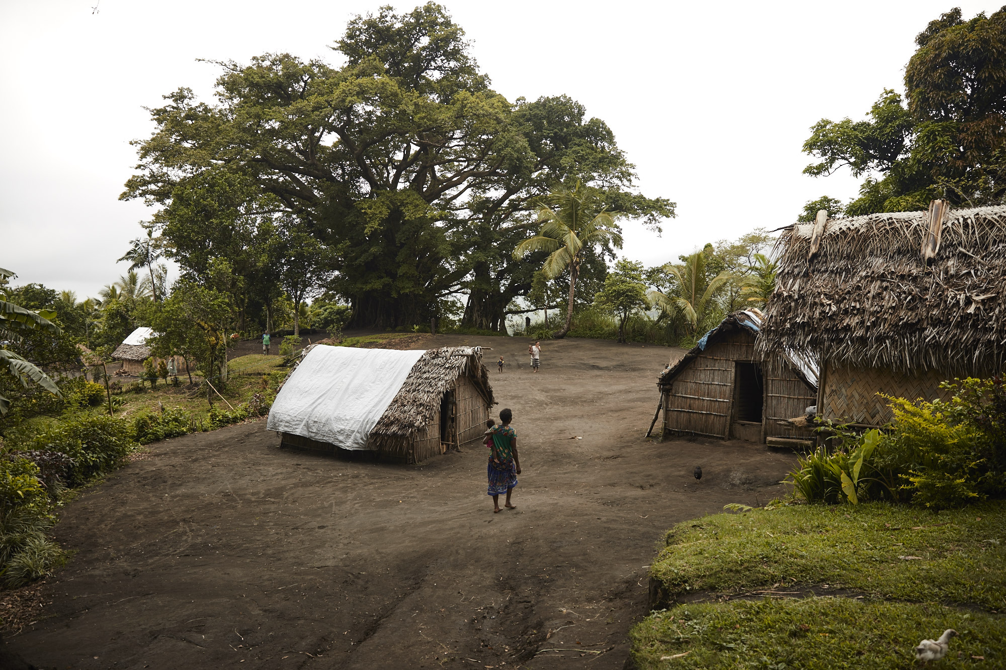 Landscape view of a small village with thatched huts, a woman carrying a child, and lush tropical trees in an outdoor setting.