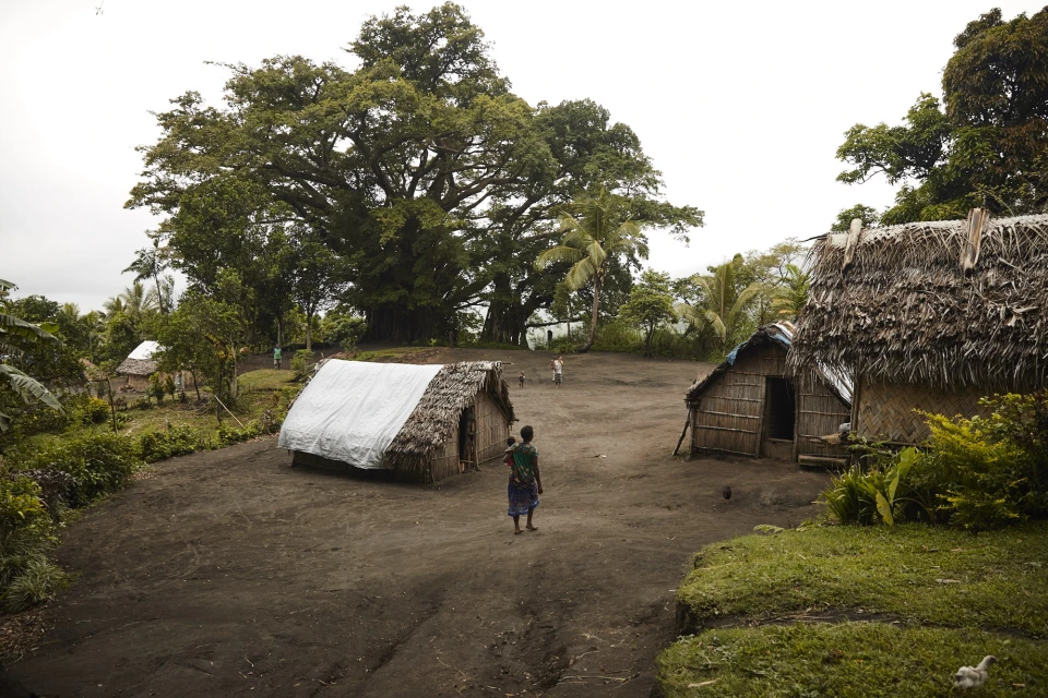 Landscape view of a small village with thatched huts, a woman carrying a child, and lush tropical trees in an outdoor setting.