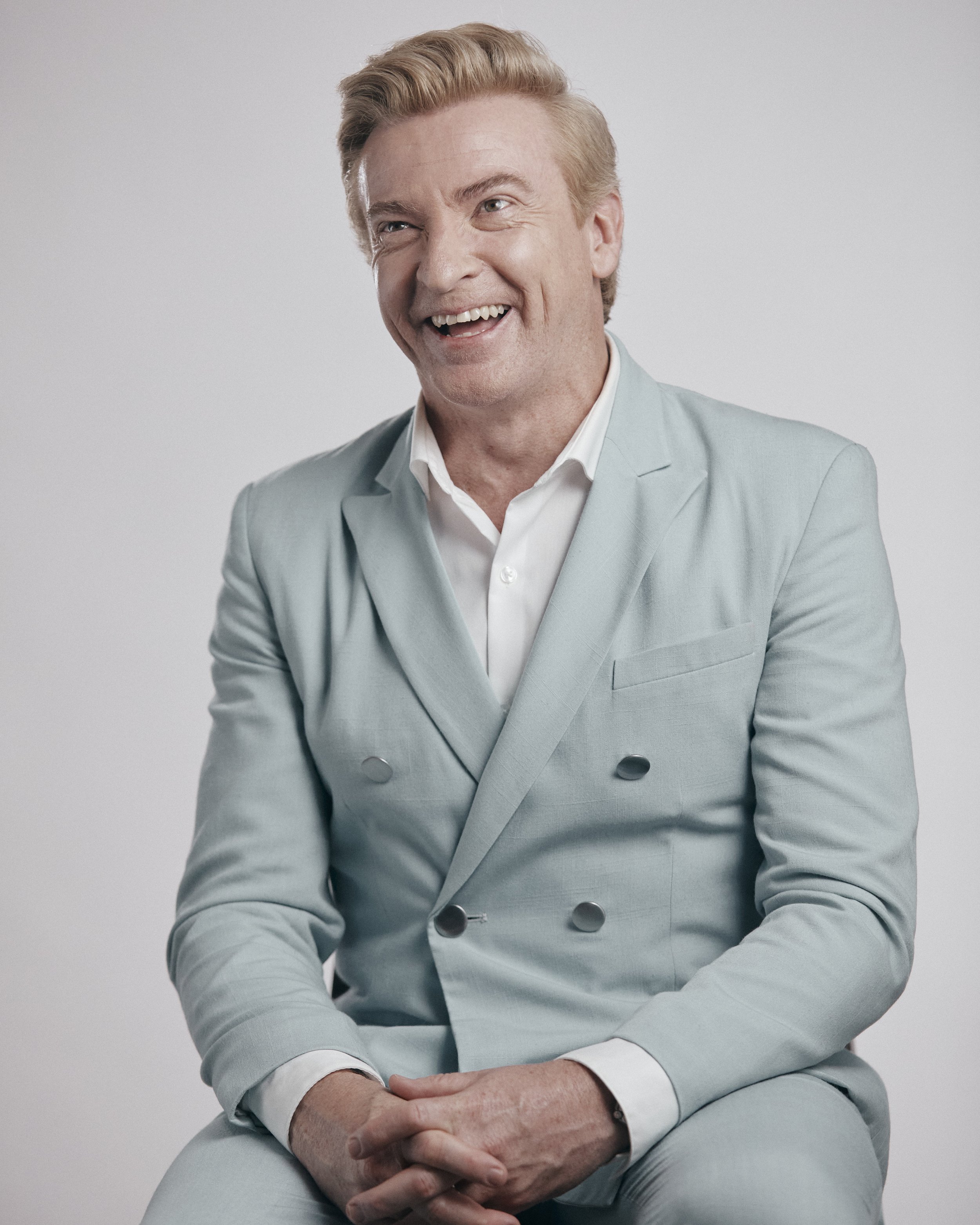 Laughing adult man in a light blue suit with a white collared shirt, sitting in a photo studio with a light background.
