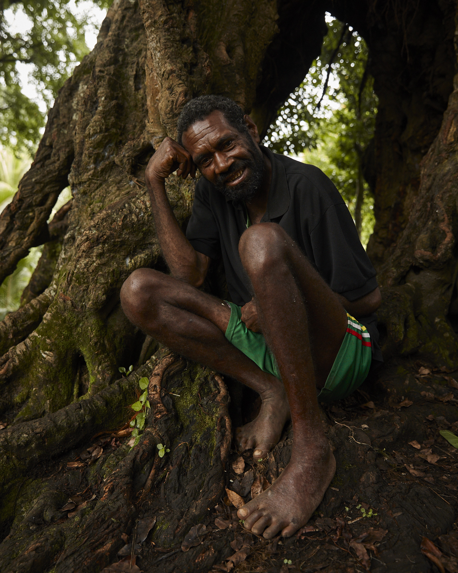 Older man with dark skin and a dark shirt sits against a large tree with exposed roots in a forest setting, smiling at the camera.
