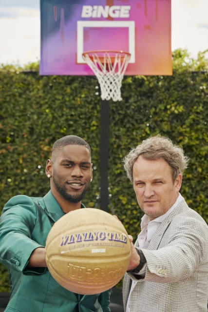 Two men hold a basketball in front of a basketball hoop with the Binge logo, set against a green leafy background in an outdoor setting.