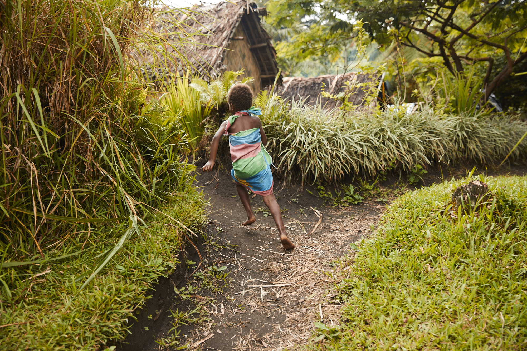 Child runs along a path near huts with thatched roofs, surrounded by lush greenery in a tropical outdoor setting on a sunny day.