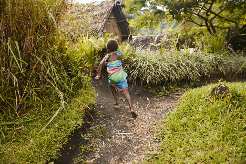 Child runs along a path near huts with thatched roofs, surrounded by lush greenery in a tropical outdoor setting on a sunny day.
