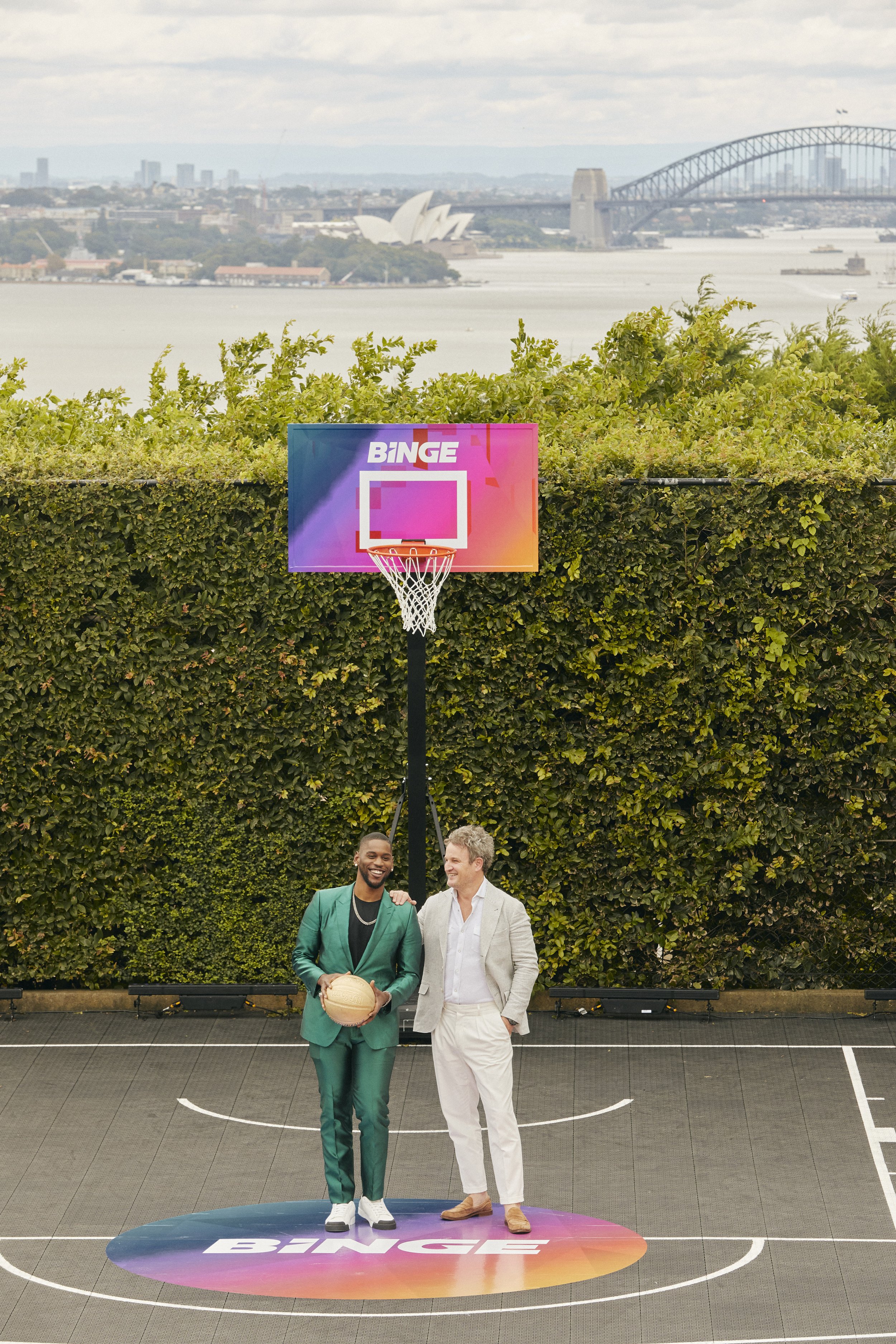 Two men pose on a basketball court with a Binge-branded hoop, set against an urban backdrop of water and city buildings.