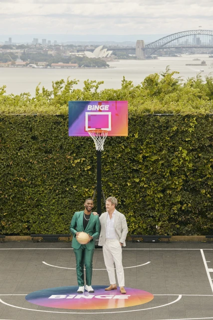 Two men pose on a basketball court with a Binge-branded hoop, set against an urban backdrop of water and city buildings.
