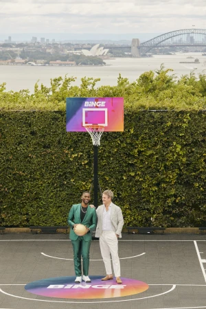 Two men pose on a basketball court with a Binge-branded hoop, set against an urban backdrop of water and city buildings.