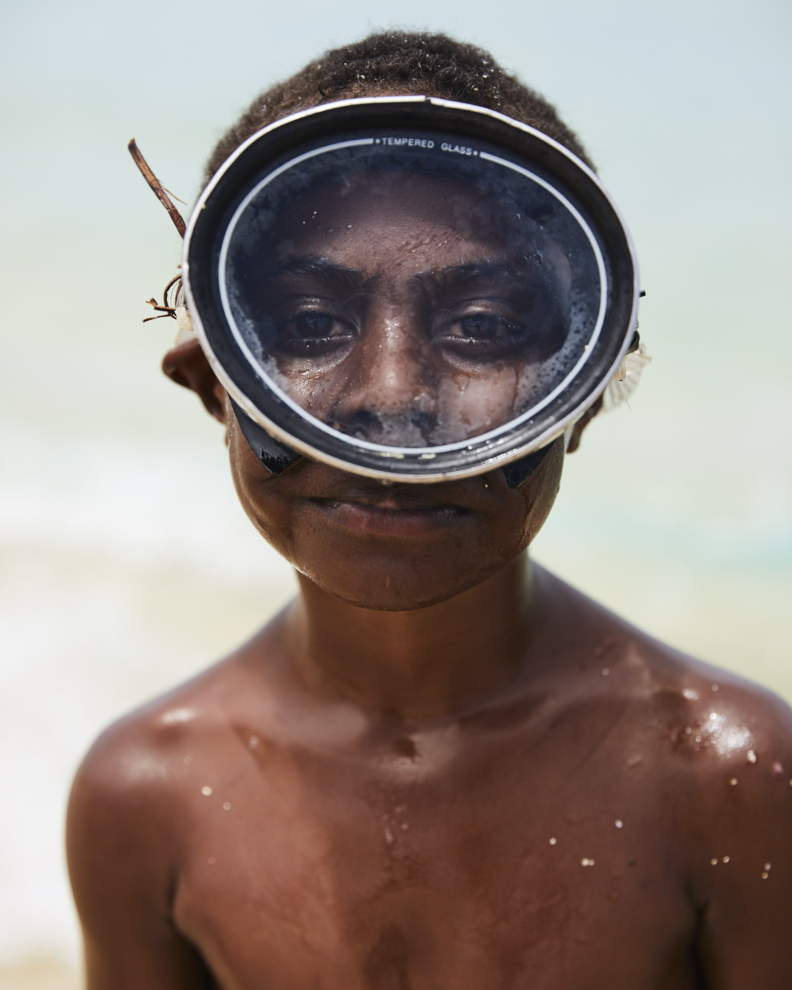 Portrait of a smiling young person with dark skin and a diving mask, standing against a blurred white beach background, close-up shot.