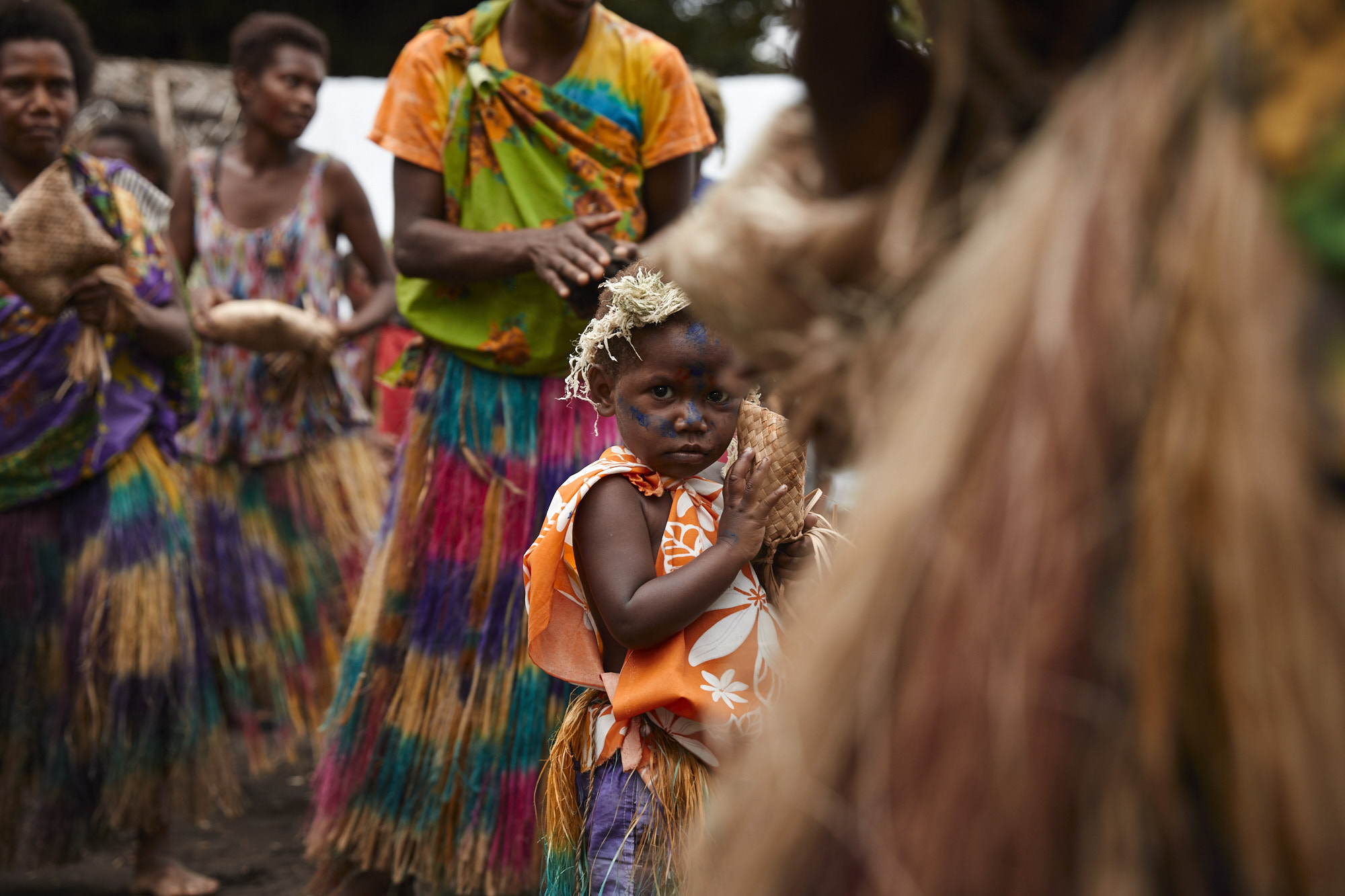 Young indigenous child with face paint holding a coconut, surrounded by a group in traditional dress with colorful skirts and tops.