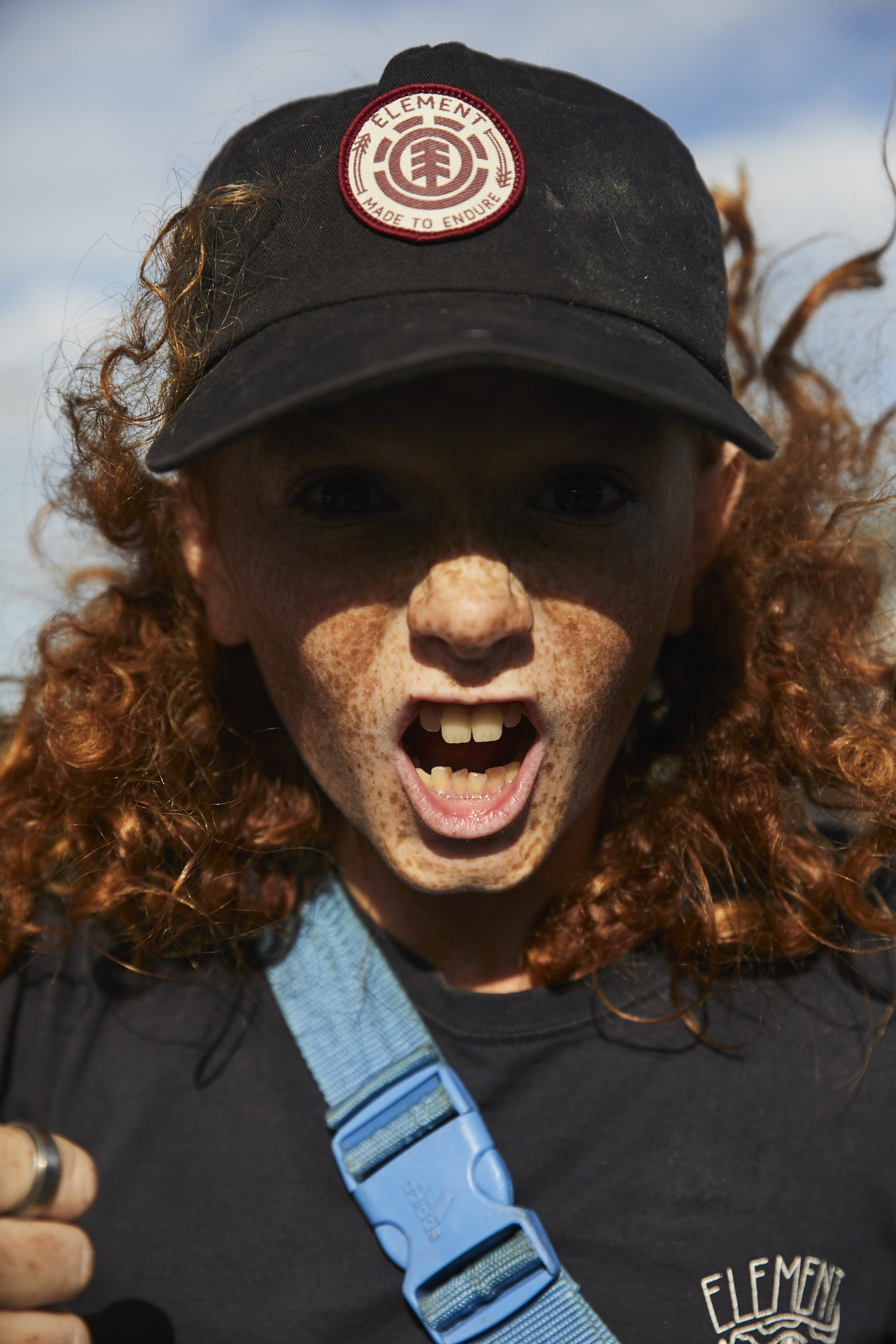 Close-up of a red-haired child in a black baseball cap with the Element logo, wearing a blue strap, and shouting with a sky background.