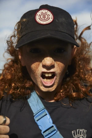Close-up portrait of a child with freckles and curly red hair wearing a black baseball cap and dark t-shirt.