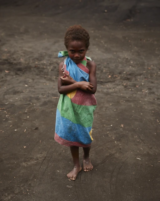 A young child stands barefoot on dark ground, draped in a colorful cloth, looking directly at the camera with a serious expression.