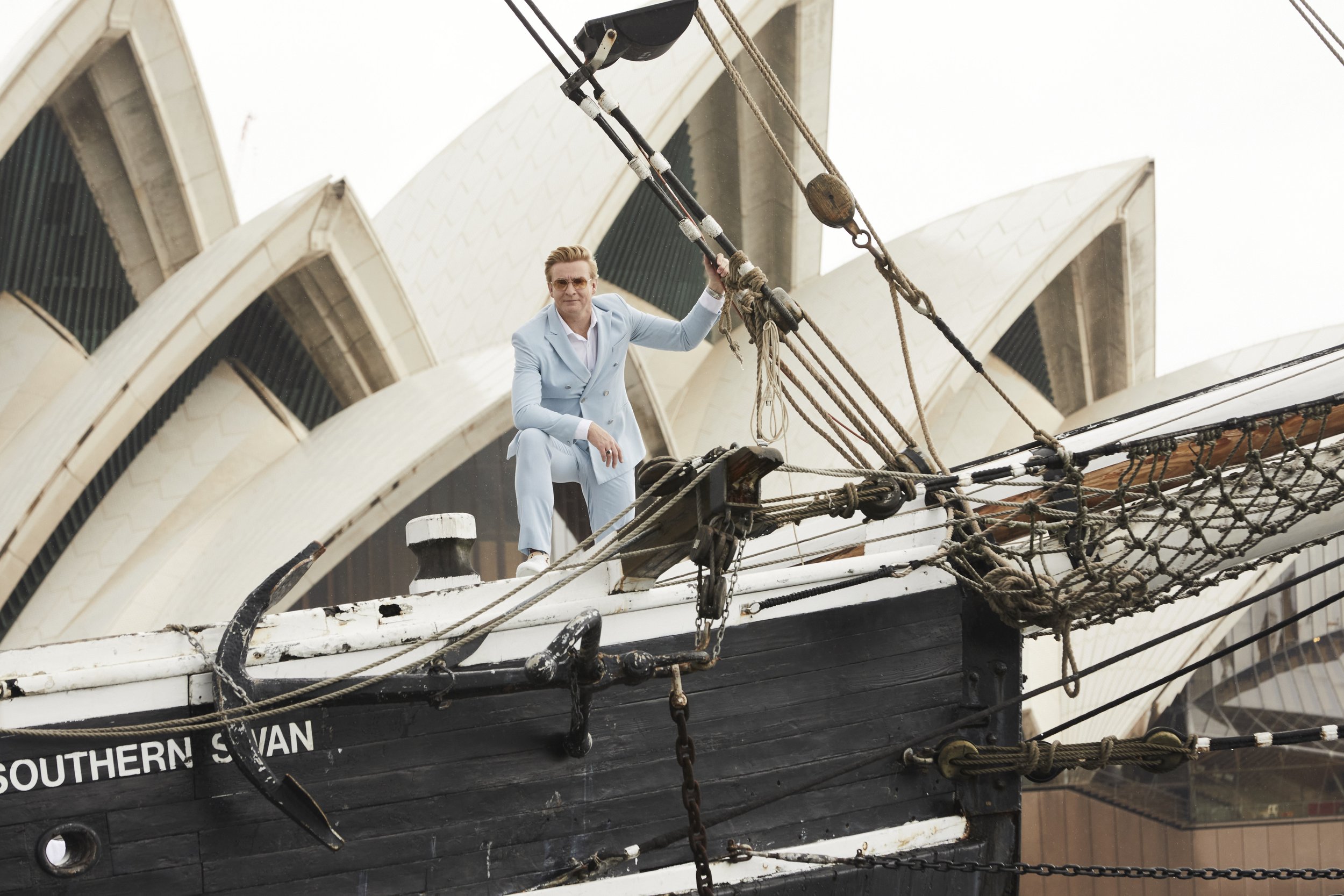 Adult male in light blue suit poses on a black ship with rigging, with Sydney Opera House in the background on a cloudy day.