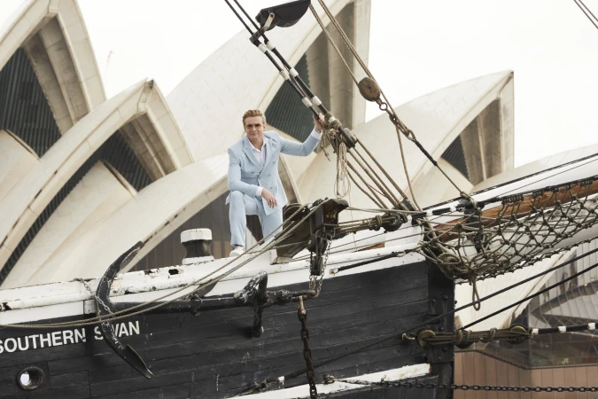 Adult male in light blue suit poses on a black ship with rigging, with Sydney Opera House in the background on a cloudy day.