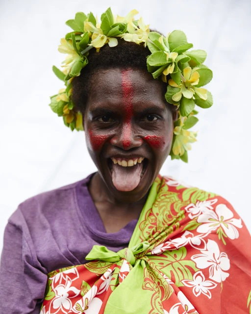 Person with floral crown and face paint, sticking out their tongue, wearing colorful patterned fabric against a plain background.