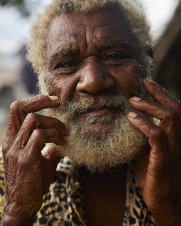 Portrait of an indigenous elderly man with salt-and-pepper beard and a leopard print shirt, holding his hands up to his face.