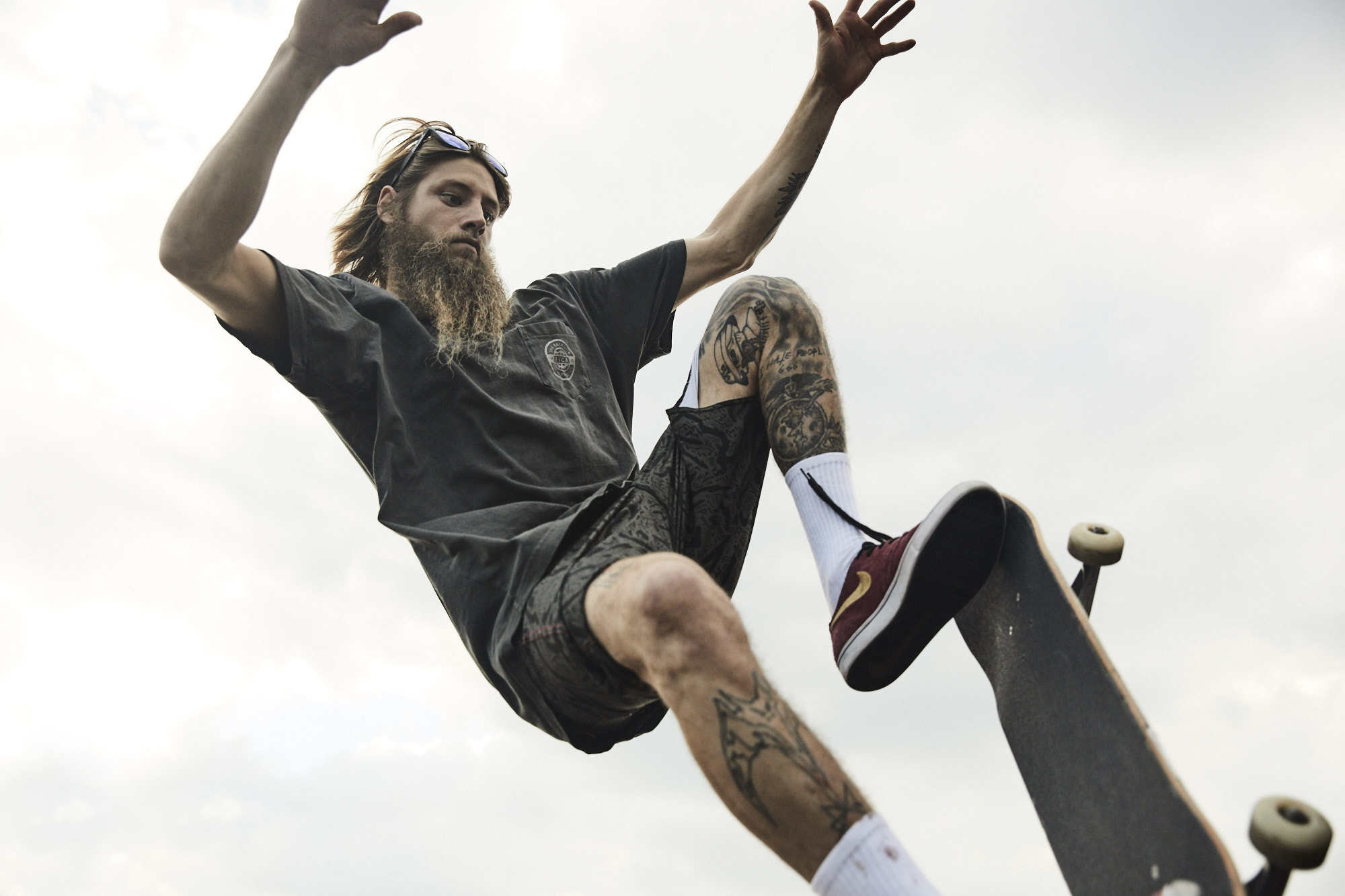 Bearded skater with long hair captured mid-air performing a trick against an overcast sky