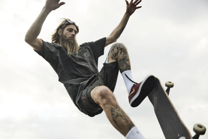 Bearded skater with long hair captured mid-air performing a trick against an overcast sky