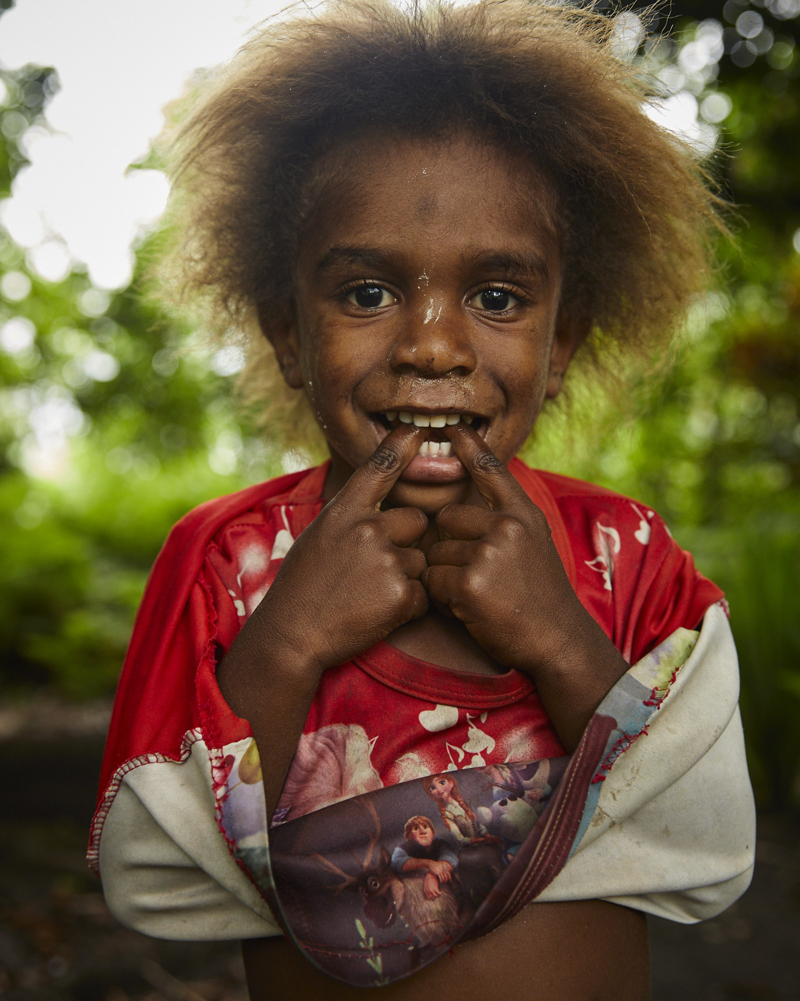 A young child with curly blonde hair smiles brightly while holding their mouth open with their fingers outdoors in a sunny, natural setting.