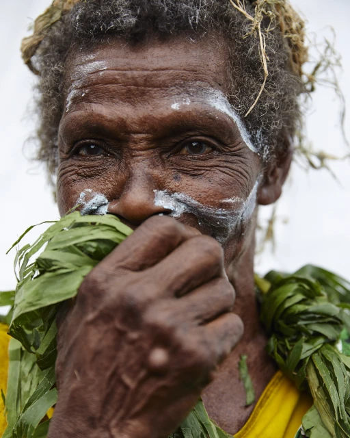 Portrait of indigenous woman with white paint facial markings, leafy necklace, and grey hair, holding leaves near her mouth.