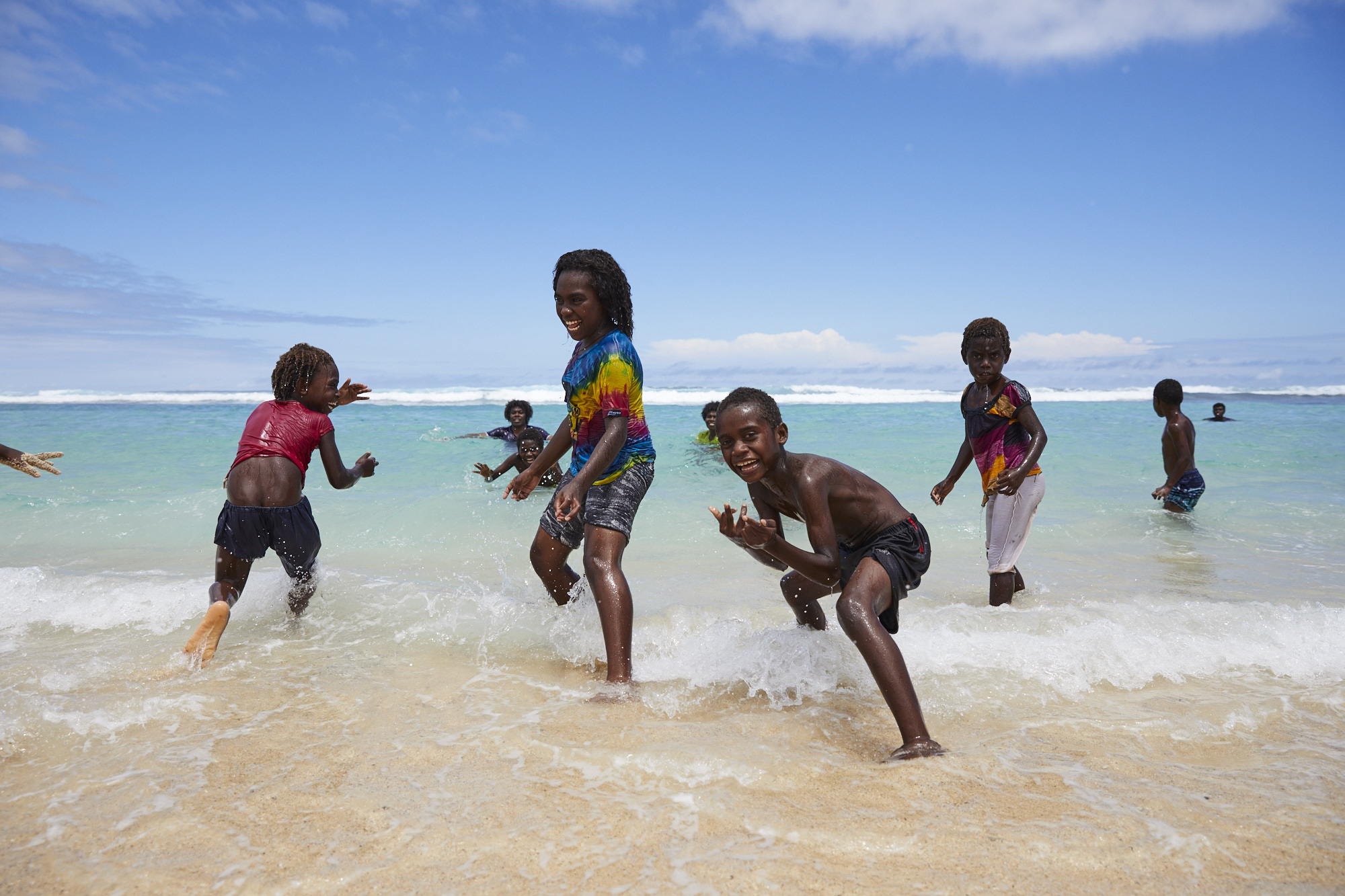 Several dark-skinned children play in the waves on a sunny beach with light blue sky, shallow depth of field, and visible horizon line.