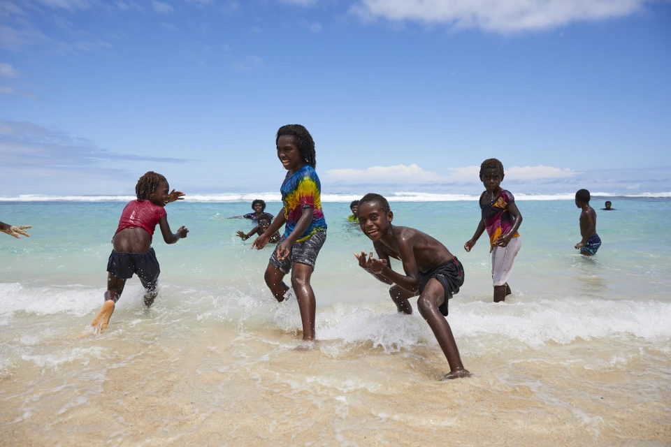 Several dark-skinned children play in the waves on a sunny beach with light blue sky, shallow depth of field, and visible horizon line.