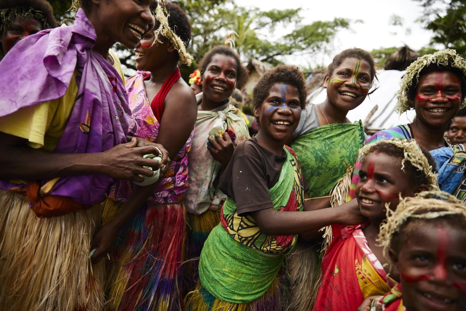 Group of smiling indigenous people with face paint and traditional clothing in an outdoor setting, showcasing cultural celebration and unity.