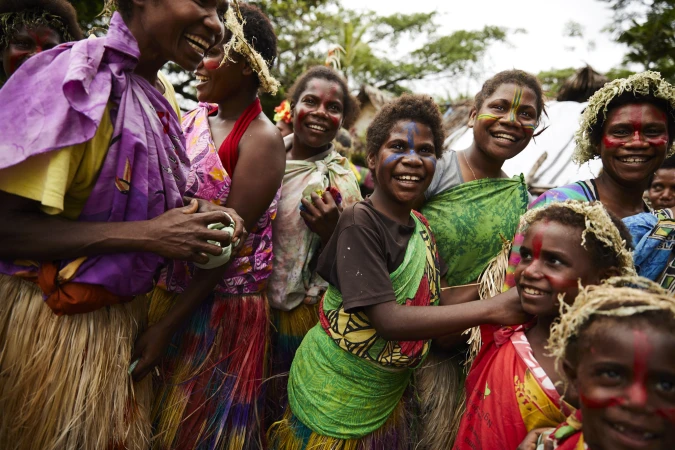 Group of smiling indigenous people with face paint and traditional clothing in an outdoor setting, showcasing cultural celebration and unity.
