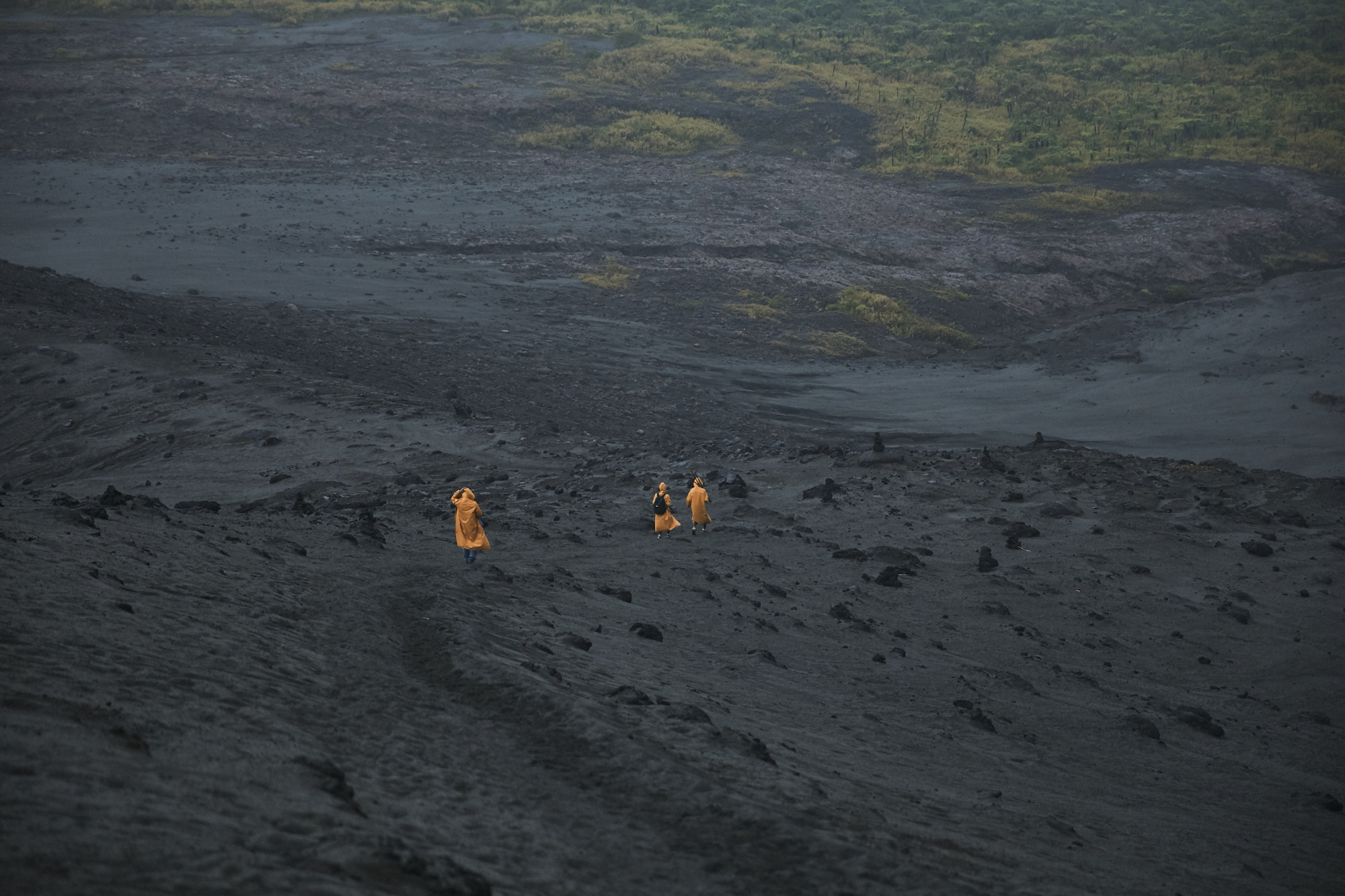 Three people in orange robes walk on a dark, volcanic landscape under an overcast sky with green vegetation in the background.