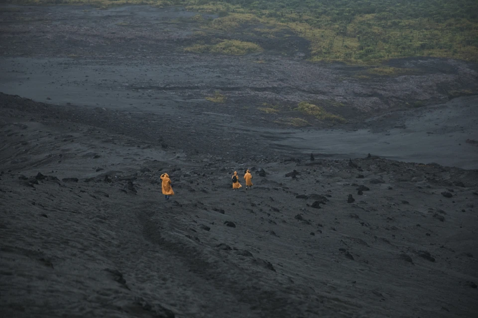 Three people in orange robes walk on a dark, volcanic landscape under an overcast sky with green vegetation in the background.