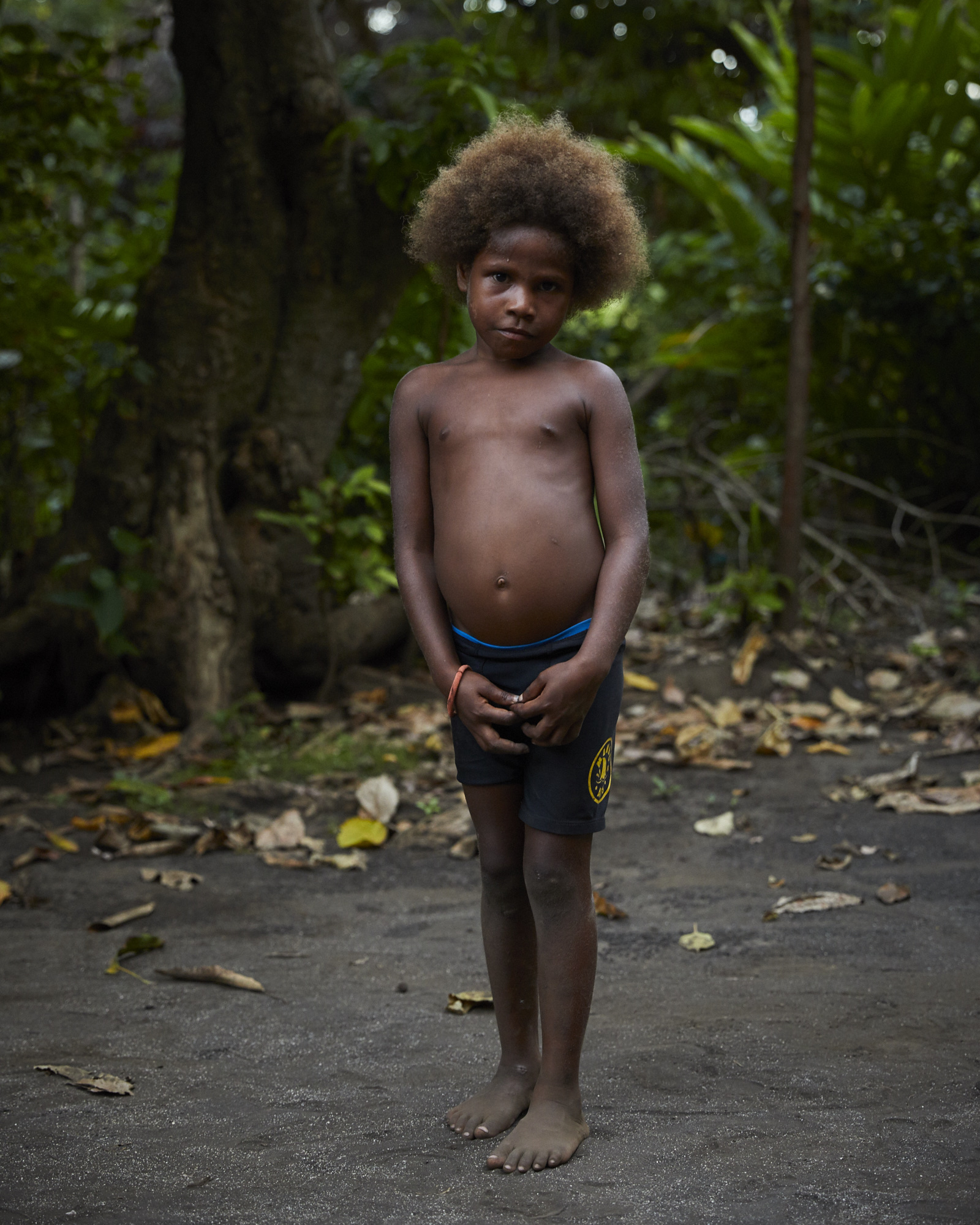Full portrait of a young child with dark skin and afro hair, standing outside in a natural environment with trees and foliage.