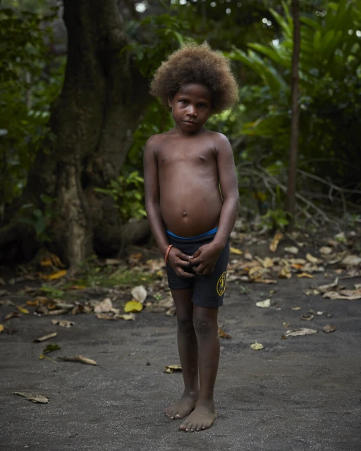 Full portrait of a young child with dark skin and afro hair, standing outside in a natural environment with trees and foliage.