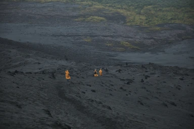 Three figures in bright yellow ponchos walking across a vast, dark volcanic landscape under an overcast sky.