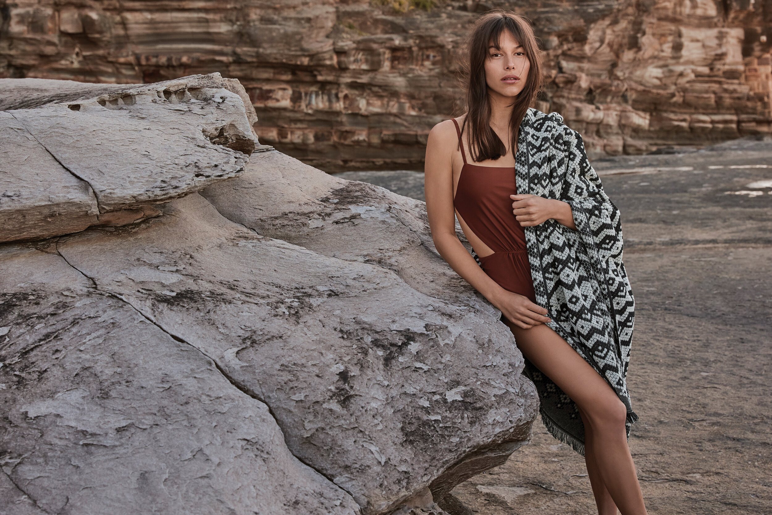 A slender woman in a brown swimsuit, wrapped in a patterned blanket, leans against a rock formation on a sunny beach.