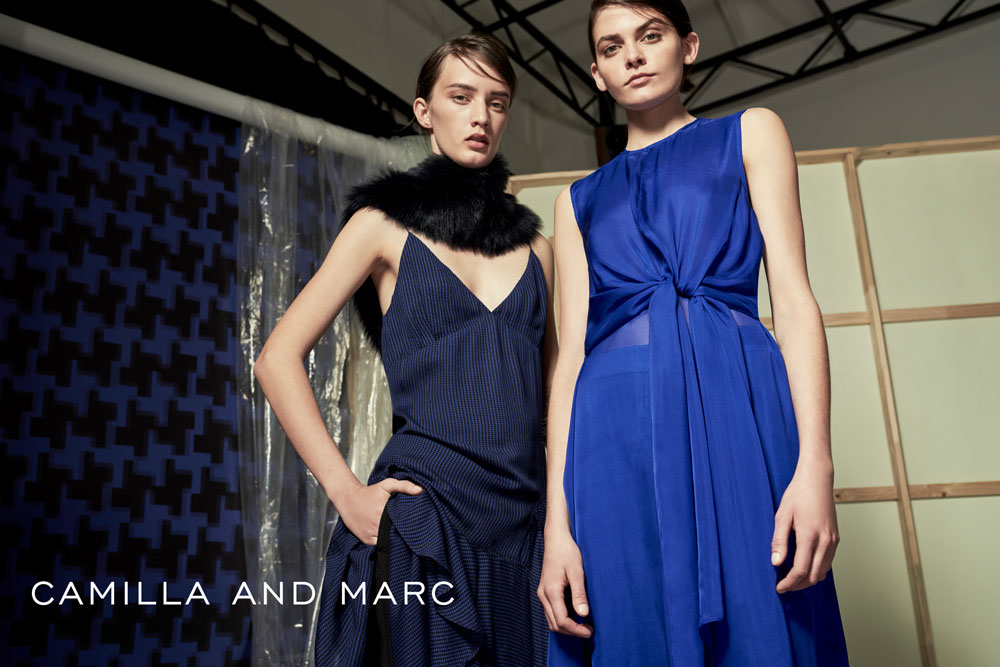 Two fashion models pose indoors in blue dresses. One model has a black fur collar. The text 'CAMILLA AND MARC' is at the bottom.