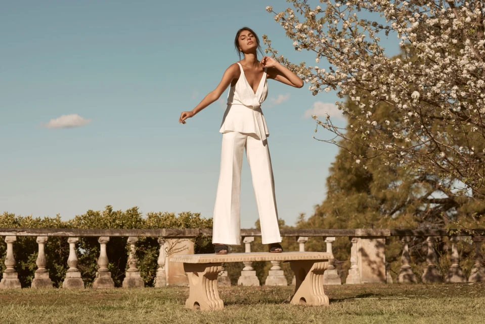 A woman in white, wide-leg pants and top stands on a stone bench outdoors on a sunny day with a blooming tree in the background.