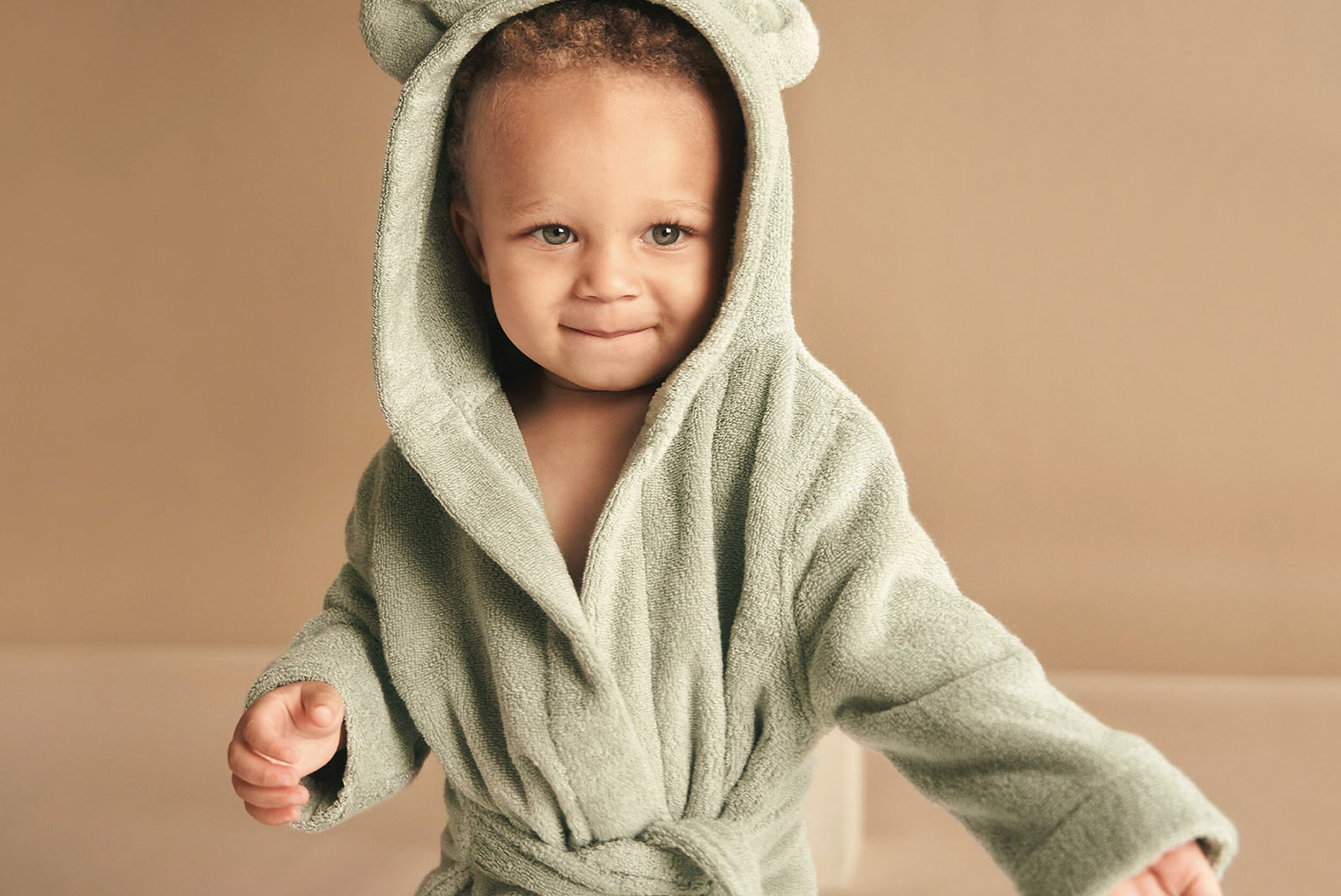 Adorable toddler with a content expression wearing a light green bathrobe with a hood and bear ears in a studio setting.