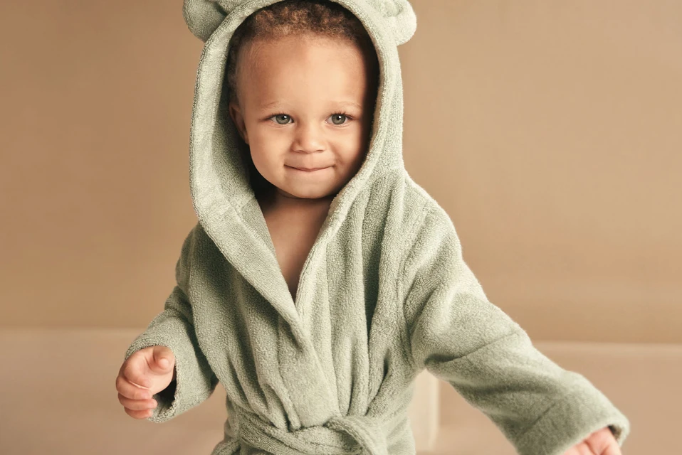 Adorable toddler with a content expression wearing a light green bathrobe with a hood and bear ears in a studio setting.