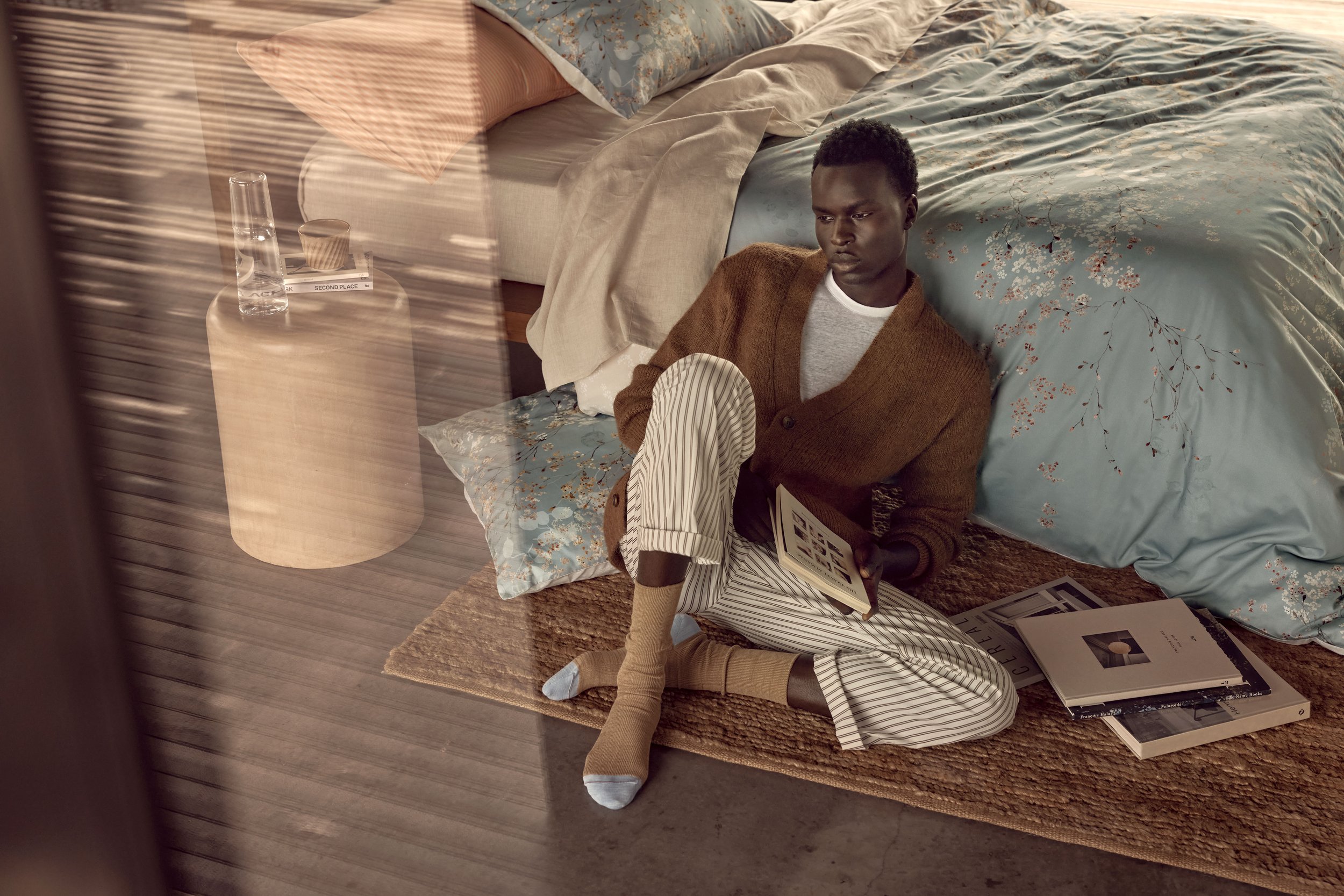 Young man relaxing indoors on a rug with a book in a striped pajama set, with a bed and a glass-topped side table in the background.