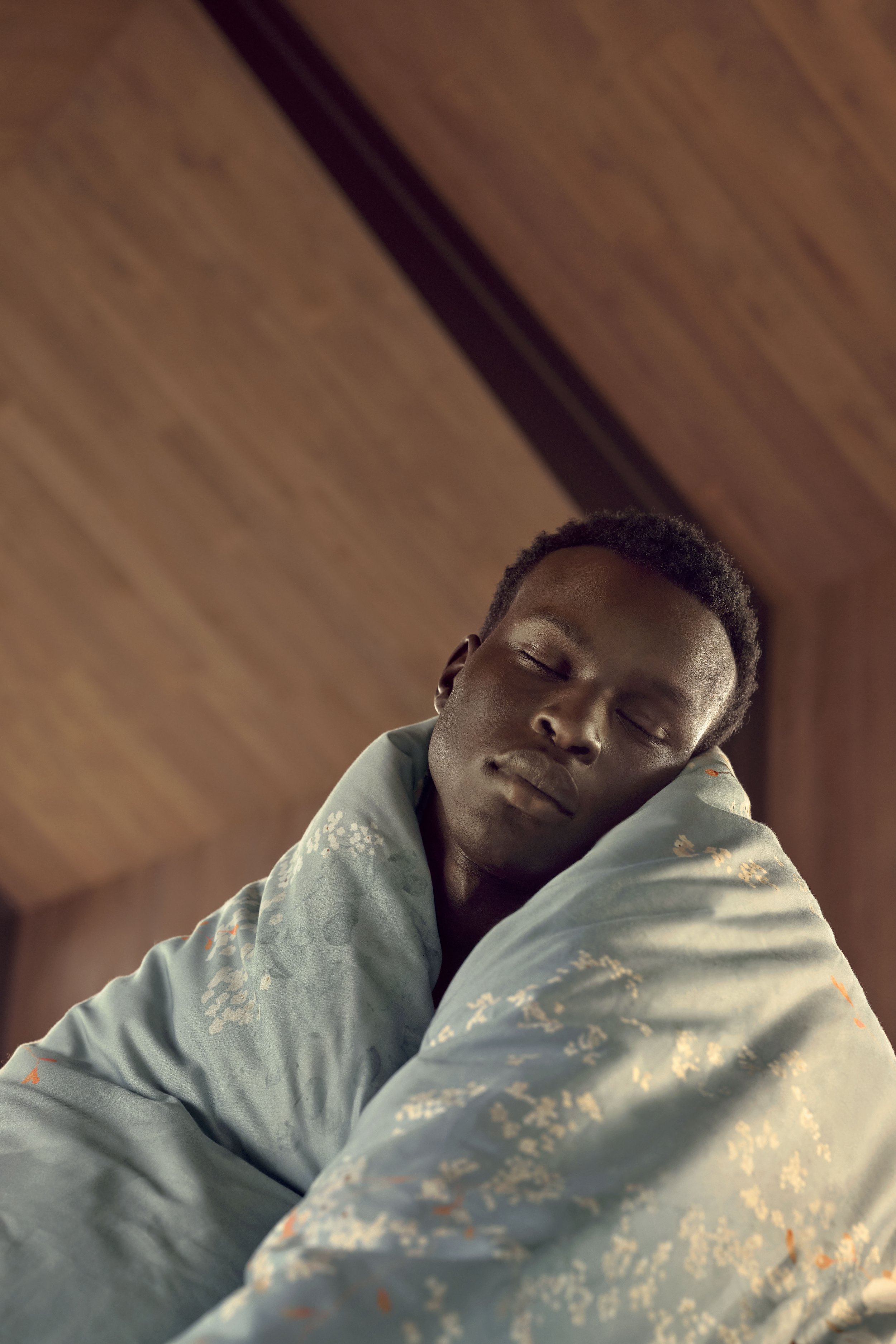 Adult male in a teal blanket with floral pattern, eyes closed, indoors against a brown wooden wall and ceiling, shot at a low angle.