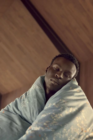 Adult male in a teal blanket with floral pattern, eyes closed, indoors against a brown wooden wall and ceiling, shot at a low angle.