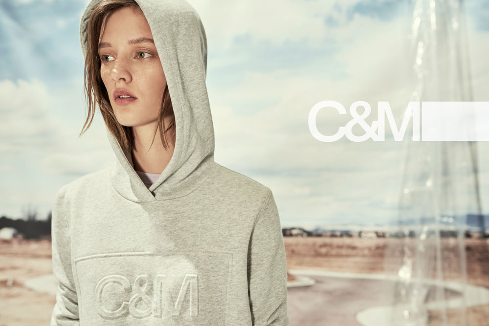Woman poses in gray hooded sweatshirt with the C&M logo against a background of a cloudy sky and stylized building.
