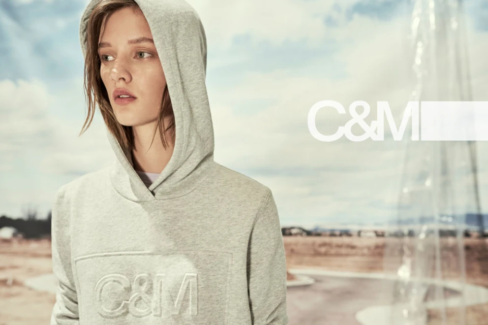 Woman poses in gray hooded sweatshirt with the C&M logo against a background of a cloudy sky and stylized building.