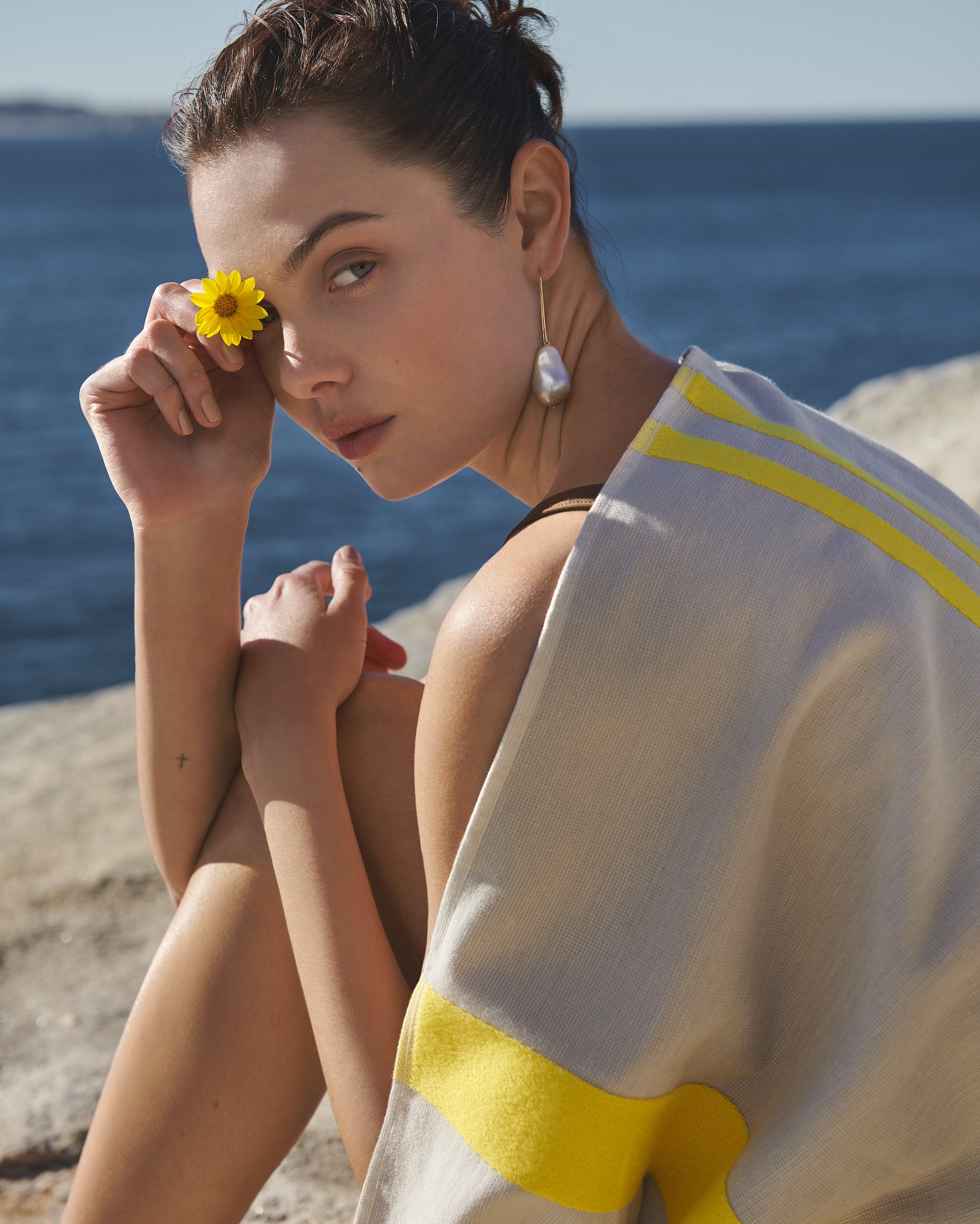 Young woman with a flower near her eye, draped in a towel with yellow stripes, sits in an outdoor setting with an ocean backdrop.