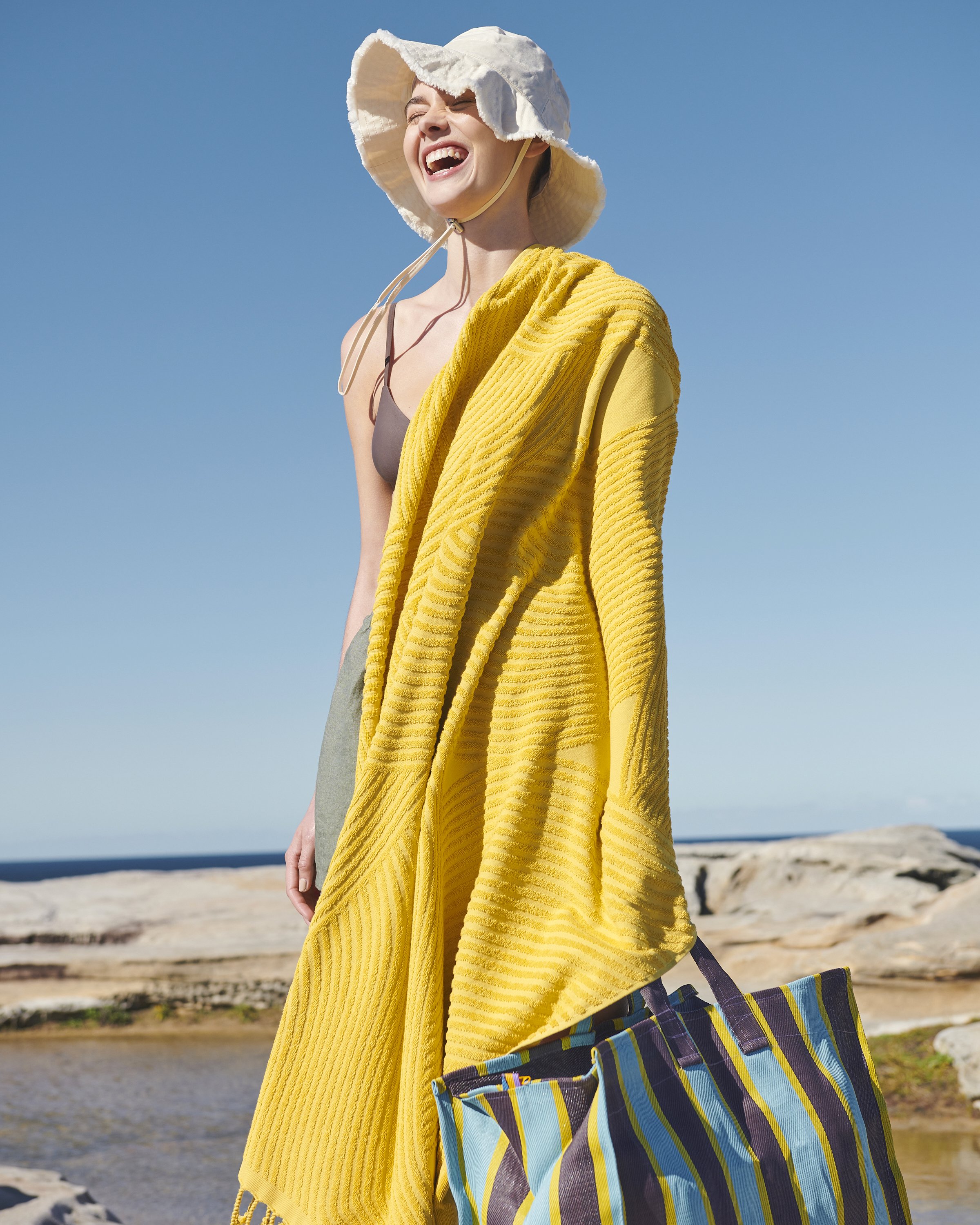 Smiling woman on rocky beach with yellow towel draped over her shoulder and blue-striped tote bag, wearing a white sun hat.